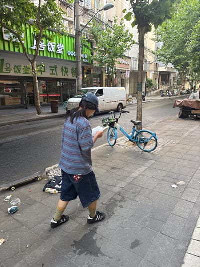 The image captures a daytime street scene in Changning District, China. A young individual walks on a grey tiled sidewalk, viewed from a rear-left angle, focused on a white rectangular object held in their hands. They wear a blue baseball cap, a horizontally striped short-sleeved shirt in blue, purple, and grey tones, dark denim shorts with a red and white patterned design on the rear pocket, yellow and blue striped socks, and black Adidas sneakers with white stripes.
To the left of the individual, a black skateboard rests on its side on the sidewalk, accompanied by a leopard-print fabric item, a dark fabric item, and a partially filled clear plastic bottle. The sidewalk exhibits numerous small pieces of debris, including cigarette butts and paper fragments, along with various stains and cracks on the grey paving slabs.
Further ahead on the sidewalk, a light blue shared bicycle is parked upright against a dark grey utility pole. The bicycle's front basket contains several green-labeled bottles. The utility pole supports multiple black overhead wires. An asphalt road runs parallel to the sidewalk. A white commercial van is parked on the street to the left. Further down the street on the right, several scooters and motorcycles are parked, and a large object covered by a brown tarp is visible at the far right.
On the left side of the street, a building features a prominent bright green sign displaying Chinese characters that translate to "No. 1 Canteen" and "Chinese Fast Food". Below this sign are glass storefronts. Multiple trees with green foliage line both sides of the street, with a large tree trunk positioned centrally. A dark brown cylindrical trash receptacle is visible near the entrance of the "No. 1 Canteen."