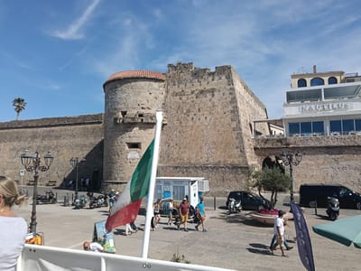 The image depicts a sunny outdoor scene in Alghero, Italy, likely along the waterfront or near a historic fortress. In the foreground, an Italian flag is prominently displayed, partially obscuring the view of several people. The background features a large, ancient stone structure, possibly a castle or fortification, with a rounded tower and crenellated battlements. A modern building with the name "NAUTILUS" in large letters is visible to the right, suggesting a commercial establishment.
Several people are walking and standing in the paved area in front of the stone structure. They are dressed casually in warm-weather clothing, indicating a pleasant day. Various vehicles, including scooters and cars, are parked or in motion. A small ticket booth labeled "TICKET OFFICE CAVE ENTRY ONLY" is also visible. The sky is blue with scattered white clouds, and a lone palm tree stands against the horizon. The overall impression is a bustling tourist area with a blend of historical architecture and modern amenities.