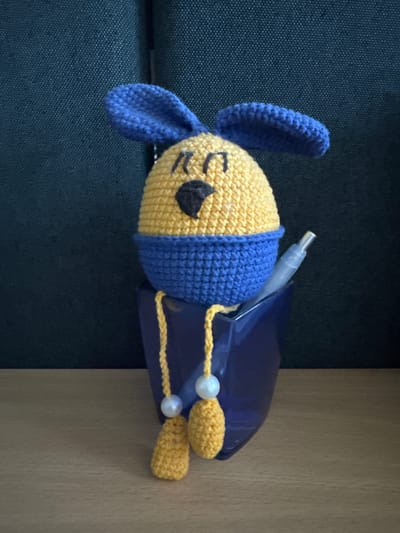 A crocheted, yellow Easter egg character with blue ears and a blue collar sits in a dark blue cup. The character has black embroidered eyes and a black embroidered nose. Two yellow crocheted legs with white beads hang from the collar, resting on the rim of the cup. A light blue pen is also placed in the cup, next to the character. The cup sits on a light brown wooden surface, with a dark textured background behind it. This scene does not appear to be located in Krakovany, Slovakia.