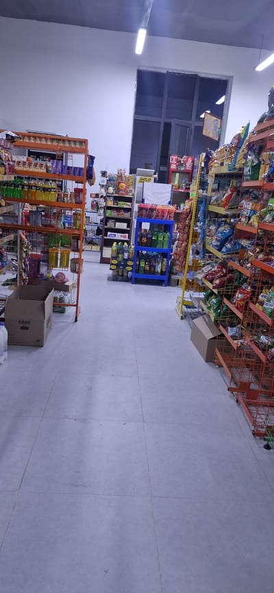 The image shows the interior of a small convenience store located in Arys, Kazakhstan. The store is stocked with various goods, including snacks, beverages, and other grocery items displayed on metal shelves. The floor is tiled with light-colored squares. In the foreground, a cardboard box and some cans are visible. In the background, there is a large window or door, and fluorescent lighting fixtures are mounted on the ceiling. The store appears to be well-lit and organized. There are no people visible in the image.