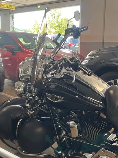 In a covered parking garage in Weiswampach, Luxembourg, a black Harley-Davidson motorcycle is parked. The motorcycle is the primary subject, positioned in the foreground and taking up most of the frame. It features a large windshield, chrome accents on the handlebars and engine, and black saddlebags. A red car is visible to the left of the motorcycle, and a dark-colored car is parked to the right. The background shows an opening to the outdoors with trees and sunlight filtering in. Above the vehicles, a yellow sign with black lettering is mounted on the ceiling, warning about "giftigung's gefahr" (poisonous danger) and urging people to leave quickly and turn off engines. The scene appears to be a typical parking area with no people present.