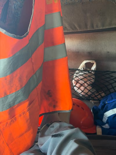 In Zarech'e, Russia, an orange high-visibility vest is prominently displayed, exhibiting dirt and wear. Behind it, an orange hard hat rests next to a blue garment with white stripes, likely workwear. A mesh net contains a white plastic bag filled with light-colored, possibly food, items. The background consists of textured brown surfaces, suggesting the interior of a vehicle or storage area.