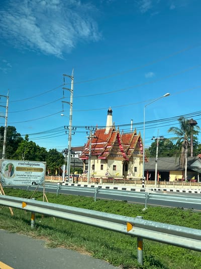 The image depicts a roadside scene in Ban Ko Pru, Thailand, under a clear blue sky with scattered clouds, suggesting it is daytime. The perspective is from a moving vehicle, indicated by the blur of the guardrail and grass in the foreground.

The main subjects are a Buddhist temple with a distinctive red and gold tiered roof and a tall, slender crematorium tower. The temple appears to be a place of worship and mourning, as evidenced by the signage in the foreground.

The visible text on the sign is a funeral announcement, reading "งานบำเพ็ญกุศลศพ คุณพ่อสน ขุนทอง (คุ้มภัยรนด์) อายุ ๙๐ ปี เริ่มงานวันที่ ๒๗ - ๒๘ มิถุนายน พ.ศ. ๒๕๖๔ ณ บ้านหนองสายแก้ว หมู่ ๔ ตำบลทุ่งโพธิ์ กำหนดฌาปนกิจวันจันทร์ที่ ๒๙ มิถุนายน พ.ศ. ๒๕๖๔ ณ เมรุวัดสำนักขัน" which translates to "Merit-making ceremony for the deceased Mr. Son Khunthong (Kumphai Rand) age 90 years. Ceremony starts on June 27-28, B.E. 2564 at Ban Nong Saikaew, Moo 4, Thung Pho Subdistrict. Cremation is scheduled for Monday, June 29, B.E. 2564 at Wat Samnak Khan crematorium." The announcement includes a portrait of the deceased, Mr. Son Khunthong.

Other elements in the scene include power lines crisscrossing the sky, a lamppost, a palm tree, and lush green vegetation along the roadside. The presence of Thai flags on the temple and the style of architecture firmly place the location in Thailand. The overall atmosphere is one of a typical day in a Thai community, with the solemnity of a funeral ceremony being announced.