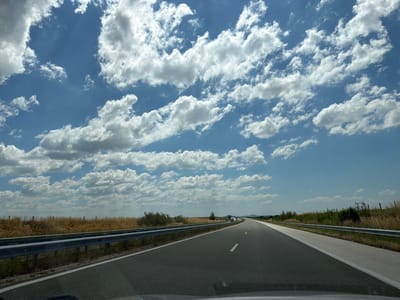 A wide, asphalt highway stretches into the distance under a vast blue sky dotted with fluffy white clouds. The road, marked with white dashed lines, curves gently to the right. A metal guardrail runs along the left side of the highway, separating it from a dry, grassy field. In the distance, a few vehicles, possibly trucks, are visible on the road. The landscape appears rural, with sparse vegetation and a fence line visible in the background. The image was taken from a moving vehicle.