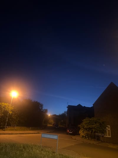 This image captures a street scene at dusk in the Borough of Swale, United Kingdom. The sky is a deep twilight blue, transitioning to black at the top of the frame. A bright street lamp illuminates the left side of the image, casting a warm glow on the surrounding trees and foliage. In the foreground, a paved road curves to the right, bordered by grass and a low wooden fence.
On the right side of the road, a brick building with a pitched roof is visible, with a window and a street sign that reads "MULBERRY WAY". Further down the street, a red car is parked. In the distance, other buildings and trees are silhouetted against the darkening sky, with additional streetlights visible. The scene appears to be a residential area with well-maintained landscaping. The image is devoid of any people or any indication of activity.