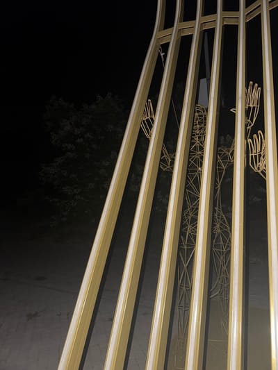 The image shows a golden structure with vertical bars and internal lighting at night. Behind the structure, there are wireframe sculptures resembling human figures, with prominent hands. A tree with illuminated blossoms is visible in the background, set against a dark sky. The ground appears to be paved. This scene is located in Egorlykskaya, Russia.