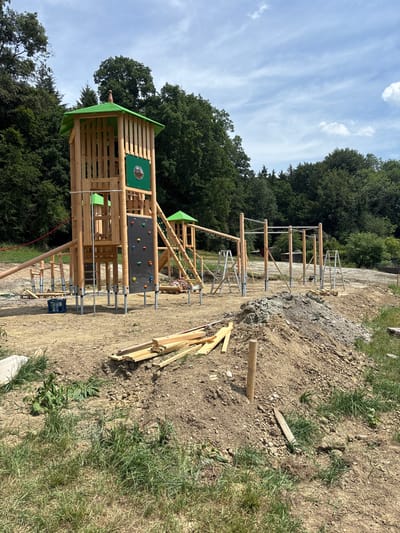 This is a daytime, eye-level, outdoor photograph of a playground under construction in the city of Buch, Germany. The main subject is a large wooden playground structure with a green roof and a climbing wall. To the right of the main structure, there are parallel bars and other wooden posts that appear to be part of another play area. The ground is covered in dirt and construction materials, including piles of wood, scaffolding, and ladders. In the background, there is a dense forest and a cloudy sky. The scene suggests that the playground is still in the early stages of development, with unfinished elements and visible construction debris.