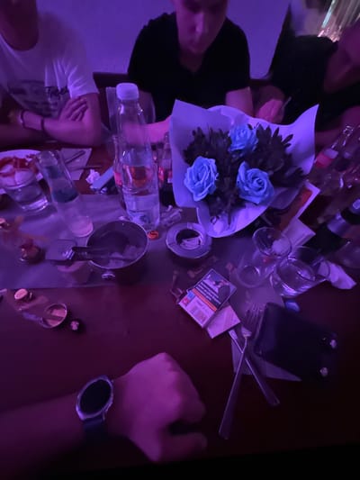 The image, apparently taken at a low angle, depicts a cluttered table in what seems to be a dimly lit establishment. The environment is marked by intense violet-blue illumination, casting an atmospheric hue.
On the table, there is a diverse array of objects, suggestive of recent activity. At the center, a bouquet of blue roses wrapped in white paper draws attention. Surrounding the flowers, we see glasses, several bottles of liquid (likely alcohol or water), and a smartphone. Also visible is a pack of cigarettes and a lighter. Cutlery – forks and knives – lie haphazardly on the table.
Fragments of people are visible around the table, indicating multiple individuals are present. A person wearing a white t-shirt is on the left side, and partially visible persons are on the top and left sides. A hand wearing a wristwatch is visible on the bottom right.
The image gives the impression of a social gathering in Panagyurski kolonii, Bulgaria, although specific details connecting it to that location are absent. The chaotic arrangement of objects and the partial presence of people suggest a spontaneous or dynamic situation.