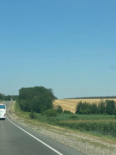 An image shows a highway with a white bus in the foreground on the left side. The bus is traveling on a road that curves to the left. To the right of the road, there is a field of dry straw and a field of corn. In the background, there are trees and a clear blue sky. The scene appears to be in a rural area.