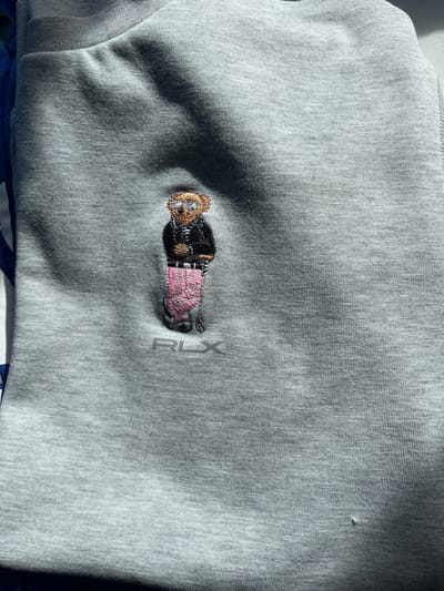 The image presents a close-up forensic view of a light grey textile garment, observed in Vieux, France. The central subject is an intricate embroidered motif located on the upper left portion of the fabric. This embroidery depicts a brown teddy bear figure, rendered wearing silver-rimmed sunglasses, a dark-colored jacket, and horizontally striped trousers in pink and white. Directly below the teddy bear, the stylized acronym "RLX" is embroidered in a metallic grey thread. The fabric itself shows subtle creasing and areas of shadow, indicating indirect or partially obstructed lighting conditions. No individuals are present in the frame, and no specific actions or interactions are depicted. The background appears to be the garment itself.