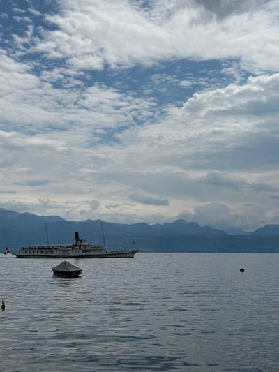 The scene depicts a calm expanse of Lake Geneva near Lausanne, Switzerland, under a sky exhibiting a mix of blue patches and scattered white and grey clouds. A multi-deck paddle steamer, white with a dark funnels and displaying a Swiss flag from its stern, traverses the water from left to right across the mid-ground. Passengers are visible on its open upper decks. In the foreground, a small, covered vessel with a dark red hull and light-colored canvas cover is moored on the water. A dark, spherical buoy floats further to the right, and a yellow cylindrical buoy is partially visible in the lower-left corner. A range of distant mountains, some with visible snow-capped peaks, forms the horizon line.