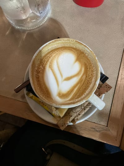 An overhead shot captures a cozy cafe scene, likely in Chaidari, Greece. The primary focus is a white ceramic cup of latte or cappuccino, adorned with intricate heart-shaped latte art on its frothy surface. Small bubbles ring the edge of the foam, indicating its freshness. The cup sits on a matching white saucer, which also holds several brown and yellow sugar packets and a metal spoon. One of the brown sugar packets clearly displays the brand name "Zazyoon."
To the left of the coffee, a clear glass contains water and multiple ice cubes, showing condensation on its outer surface, suggesting a cool refreshment. A portion of a red, possibly plastic or ceramic, object is partially visible on the far left. The beverages are placed on a light-colored, textured tabletop, which is bordered by a wooden frame on the top and right sides. In the bottom right corner, a dark-colored object, possibly a purse or bag, is visible, adding a hint of a person's presence just outside the frame. The lighting appears soft and warm, typical of an indoor cafe setting, without direct sunlight, making the specific time of day ambiguous. The overall image evokes a sense of relaxation and enjoyment of a coffee break.