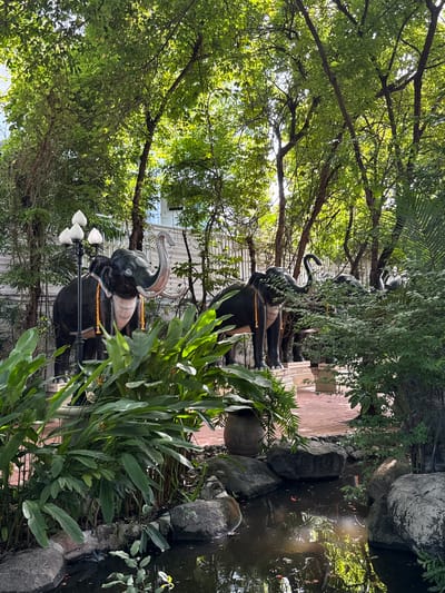 The image captures a scene in Samrong Nuea Subdistrict Municipality, Thailand, featuring a row of elephant statues lining a pathway. The statues are black and appear to be adorned with yellow garlands. Lush green vegetation surrounds the statues, including large-leafed plants in the foreground and trees that create a canopy overhead. A body of water is visible to the right, with rocks lining its edge. In the background, a white building can be partially seen through the trees. The overall impression is that of a tranquil garden or park-like setting. A lamppost with white globe lights stands next to one of the elephant statues.