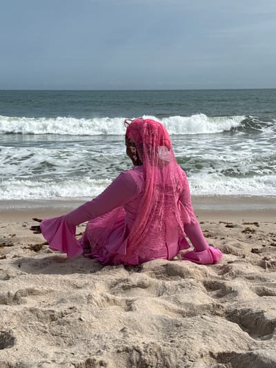 A person dressed in a pink garment kneels on a sandy beach. The ocean is visible in the background with waves crashing. The location is Mopo-Akinlade, Nigeria.