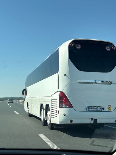 A white Neoplan bus is traveling on a highway on a clear, sunny day. The bus has tinted windows and speed limit signs indicating 80 and 100. A white car is visible in the distance on the same highway. The bus has a license plate that reads "PP 9625 AH". The environment appears to be a rural highway, likely in Serbia, given the license plate and the overall landscape. The image is taken from another vehicle traveling behind the bus.