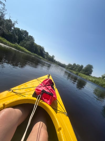 The image shows a first-person perspective from a yellow kayak on a wide, dark river. The kayak's bow is visible in the foreground, with a red life vest strapped to it. A person's legs are visible inside the kayak, with some water droplets on their skin. The riverbanks are lined with dense green trees and tall grass. The sky is a clear, bright blue with no clouds. The overall scene suggests a peaceful, sunny day spent kayaking on a natural waterway. The location is identified as the city of Kurmene, Latvia.