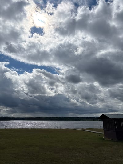 The image depicts a landscape scene, likely on a cloudy day, with a body of water in the mid-ground and a grassy area in the foreground. The sky is filled with dramatic, dark, and light gray clouds, with patches of blue visible. The sun is partially obscured by the clouds, creating a bright, hazy effect.
On the left side of the image, a silhouetted figure is walking across the grassy area, carrying a load on their shoulder. Further out on the water, there are a few small, indistinct figures, possibly people or objects. A wooden structure, resembling a small shed or shelter with a dark roof and open sides, is situated on the right side of the foreground. A wooden walkway leads from the grassy area towards the water, near the shelter. The water appears somewhat choppy, reflecting the sky. A distant treeline forms the horizon behind the water.
While no specific landmarks are visible to pinpoint the exact location, the description of the landscape suggests a natural or recreational area, possibly a park or a lakeside resort. The presence of a body of water, grassy areas, and a shelter points to a place where people might gather for outdoor activities. Without further information or distinct visual cues, it is impossible to definitively identify the location within Мазалаўскі сельскі Савет, Belarus.