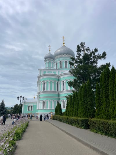 This is a ground-level, vertical, eye-level shot of the Holy Trinity Cathedral in Diveyevo, Russia, on an overcast day. The cathedral is a prominent, large, light turquoise structure with white trim and silver domes topped with golden crosses. It features multiple arched windows and decorative columns. To the right of the cathedral, a row of tall, dark green cypress trees lines a manicured hedge. In the foreground, a paved walkway curves to the right, bordered on the left by a flower bed with light purple blooms and dense green foliage. A paved path leads away from the viewer toward the cathedral. People, dressed in modest attire, are walking along the path and gathered in front of the cathedral. A lamppost with three lights stands to the left of the path. The overall atmosphere is serene and spiritual.