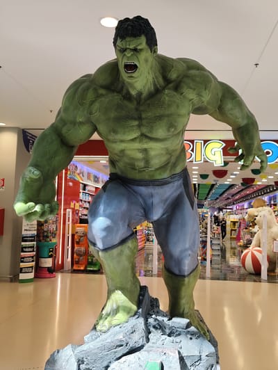 A full-body statue of the Hulk, a fictional green-skinned superhero, stands prominently on a grey rocky base. The statue is positioned in what appears to be a toy store, identified by signage that reads "BIG TOYS" in colorful letters. The Hulk is depicted in a muscular, enraged pose, with its mouth open and fists clenched. It wears light blue shorts. In the background, shelves are filled with toys and other merchandise, and several people are visible browsing in the aisles. To the right of the Hulk statue, a large stuffed bear and a red and white ball are also visible. The lighting suggests an indoor environment, possibly a shopping mall or a large retail store. The location is specified as the Big Toys store in Nola, Italy.