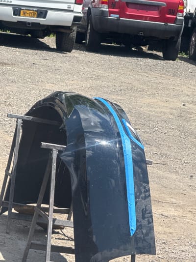 A black car bumper is supported by two metal stands on a gravel lot. The bumper has blue tape on its surface, indicating it is being prepped for repair or painting. In the background, the rear ends of two vehicles are visible: a white pickup truck with license plate JNY-7539 and a red SUV with "NY" decals. The scene appears to be outdoors in an industrial or automotive repair area.