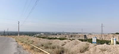 The image captures a panoramic view of the outskirts of Margilan City, Uzbekistan, under a clear, hazy blue sky. The foreground is dominated by a dirt road on the left, lined with dry, sparse vegetation and piles of sandy soil. A yellow pipe runs along the ground.

In the mid-ground, a large, excavated area or quarry reveals layered rock formations. Dense green trees are visible within the quarry, suggesting a pocket of vegetation. Several industrial structures are present, including two tall, cylindrical silos with blue and white markings, and another two silos with yellow and white markings on the right side of the quarry. A high-voltage power line pylon stands prominently to the right of the silos.

The background features a sprawling cityscape of Margilan City, with buildings of various sizes and styles spread across the horizon. More power lines stretch across the landscape, supported by pylons of different designs, including a lattice tower to the left and a more modern, slender tower further into the city. A plume of dust or smoke rises from a point within the city, possibly indicating industrial activity or construction. The overall impression is one of a semi-arid environment on the edge of an urban area, with evidence of infrastructure and industrial presence.