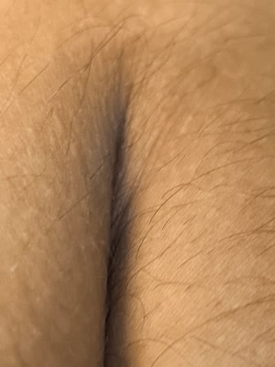 This is a close-up, extreme macro photograph of human skin, likely from an arm or leg, given the presence of fine hairs. The skin tone is light brown with some visible pores and slight variations in pigmentation, appearing as lighter speckles. The lighting creates a shadow that emphasizes a subtle crease or fold in the skin, adding a sense of three-dimensionality to the texture. The hairs are dark and sparse, scattered across the surface. The image is abstract and focuses entirely on the surface details of the skin, with no discernible objects, scene, or setting beyond the immediate texture. There is no visible text, and the image does not provide cues about the time of day, weather, location, or emotions. The context of Ново-Талицы, Russia, is not visually represented in the photograph itself.