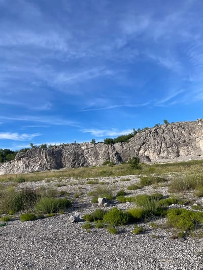 The image depicts a quarry in the city of Vrela, Montenegro. A large, layered rock face dominates the mid-ground, characterized by rough, weathered textures and varying shades of grey and white. The top of the quarry is lined with scattered green trees and shrubs. In the foreground, a gravelly expanse is dotted with patches of dry grass, small green bushes, and a few scattered rocks. The sky above is a vibrant blue, streaked with wispy white clouds. The overall scene suggests a dry, possibly arid environment under a clear sky. There are no people or animals visible in the image.