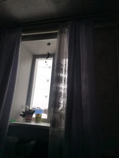A window partially obscured by grey and white curtains. A star and moon mobile hangs in front of the glass. Two potted plants sit on the windowsill. Outside, snow and houses are visible. The room is dark, illuminated only by the light filtering through the window. The overall tone is melancholic.