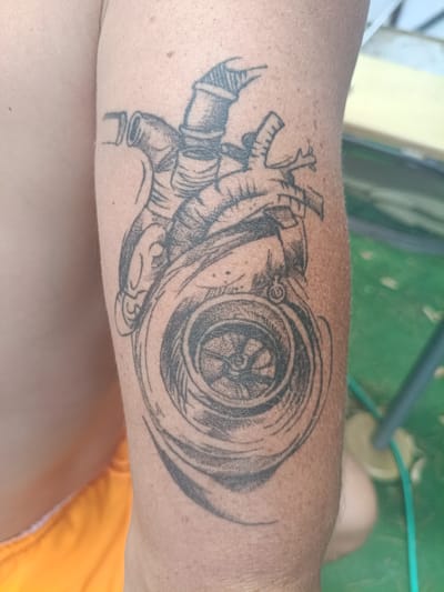 The image shows a tattoo on a person's arm. The tattoo depicts a mechanical heart integrated with a turbocharger. The style is black and grey, with heavy linework and shading. The person is wearing orange shorts, suggesting an outdoor setting. The background is out of focus but appears to be a green surface, possibly grass or a mat, with some blue and yellow elements visible. The image is cropped to focus on the tattoo and the arm. The tattoo is located on the upper arm, extending towards the elbow. The lighting suggests it is daytime.