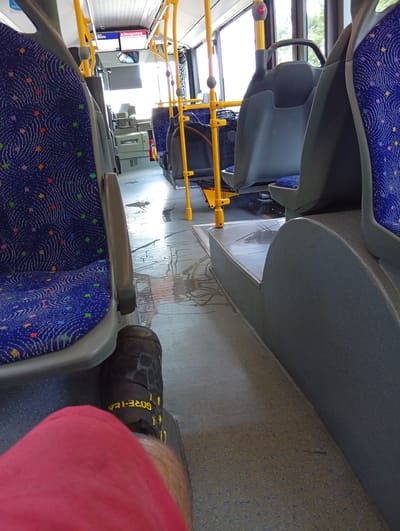The image shows the interior of a bus. In the foreground, a person's leg wearing a red garment and a black sock with yellow "GORE-TEX" lettering is visible. The bus seats are blue with a colorful speckled pattern. The aisle floor is wet and appears to have liquid spilled on it, with reflections visible. Yellow poles are a prominent feature of the bus interior. In the background, there are more seats and windows, suggesting it is daytime. One screen above the driver's area displays "Störungsmeldung" (Malfunction report) and "Bleibe" (Stay). The scene depicts a mundane moment inside public transportation, with the wet floor being a notable detail.