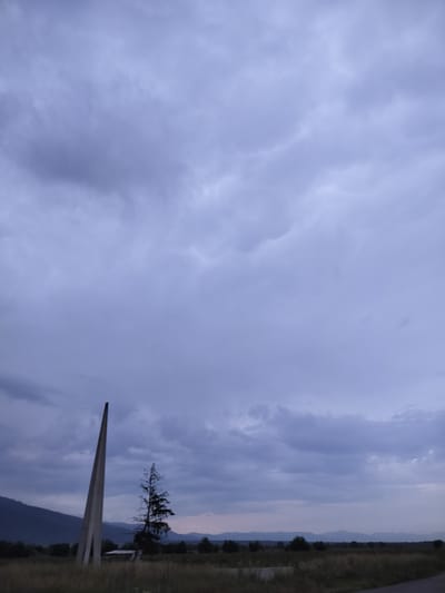 This image depicts an outdoor landscape in the vicinity of Tazha, Bulgaria, under an overcast sky. The sky occupies approximately two-thirds of the frame and is dominated by dense, grey-blue cloud formations with varying light and shadow, suggesting either an approaching storm, dusk, or dawn.
In the foreground, a field of dry, brownish-green grasses and low vegetation is visible. A dark, narrow paved road or track angles into the lower right corner of the frame.
A prominent, tall, slender monument, possibly constructed from light-colored concrete or stone, stands in the left mid-ground. It has a triangular or wedge-like cross-section and tapers to a point at its apex, angled slightly upwards and to the left. To the right of this monument, a tall, bare or sparsely leaved tree with dark, gnarled branches is silhouetted against the sky. Slightly behind and below this tree, a small, low-lying structure with a light-colored roof is discernible within the field.
The background features a horizontal band of darker green trees and bushes. Beyond this, a series of distant mountain ranges stretches across the horizon, appearing as muted, hazy silhouettes in shades of dark blue and purple. The mountains on the far left are taller and more defined. No individuals or animals are present in the scene.