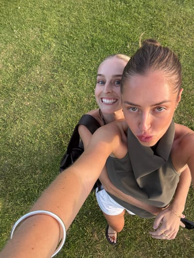 Two women pose for a selfie, likely outside, on a grassy lawn. One woman is fair-skinned with blonde hair pulled back and wearing a black top. She smiles broadly. The other woman is positioned behind and above, her light brown hair in a bun. She wears a green sleeveless top and white shorts. The selfie extends to include the arm holding the phone, featuring a silver bangle bracelet, and a glimpse of a black sandal on the left side of the frame.