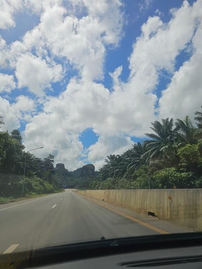 The image shows a road in Ban Nai Sa, Thailand. The road is asphalt and has white lane markings. On the left side of the road, there are palm trees and lush green vegetation, with steep, rocky hills in the background. The sky is blue with scattered white clouds, suggesting it is daytime and the weather is fair. The right side of the road is bordered by a concrete barrier. The image is taken from a moving vehicle, as indicated by the dark vertical line on the right edge of the frame, which appears to be part of the vehicle's interior or frame. There is no visible text in the image. The scene evokes a sense of travel and a tropical landscape.