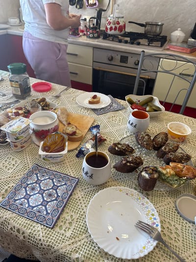 The image depicts a domestic kitchen and dining area in Klementyevo, Russia, with a table laden with food in the foreground and a person standing at a kitchen counter in the background.
In the foreground, a table covered with a light-colored, crocheted lace-patterned tablecloth displays a variety of food items and dishware. These include a large glass jar of instant coffee, a white rectangular dish with small pastries, two plastic containers of "Савушкин" (Savushkin) brand yogurt/dessert, and a white decorative bowl with red floral patterns holding a granular white substance, likely sugar. A small wooden cutting board features a piece of bread topped with sliced fruit and a glaze. A white mug with a spoon contains a dark liquid (coffee or tea), positioned near a blue and white snack wrapper. A clear, patterned glass platter holds approximately 8-10 individual dark-colored pastries, some topped with nuts, seeds, or frosting. A small white plate with a single pastry topped with a raspberry rests on a blue and white patterned coaster. A white, scallop-edged bowl contains gherkins and red peppers. An empty white plate with a colorful geometric rim pattern, containing crumbs, has a silver fork resting on it. A square blue and white patterned mat and a small empty orange-rimmed cup are also visible on the table.
In the background, a person, partially visible from the back, wears a white t-shirt and light purple pants. They stand facing a light-colored kitchen countertop equipped with a stainless steel sink, light-colored cabinets with silver handles, and a stainless steel oven and gas stovetop. Two decorative kettles, one large white with red poppy flowers and one smaller black with floral patterns, are on the stovetop/counter, alongside a metal pot on the stovetop. A metal-framed chair with a decorative backrest is positioned to the right of the table, and red flooring is visible underneath. The scene suggests a prepared meal or snack spread in a casual home environment.