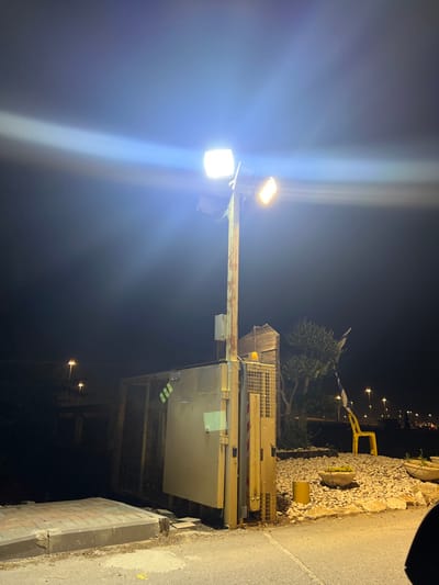 The image shows a streetlight with two lights shining brightly against a dark night sky. The streetlight is mounted on a rusted metal pole and is located in Gan HaDarom, Israel, at coordinates 31.811, 34.702. The streetlight is positioned near a gate and a large metal box. The gate is made of metal bars and is partially open. The metal box has a yellow surface and is attached to the pole of the streetlight. The image is taken from a slightly elevated position, looking down at the streetlight and the gate. The area surrounding the streetlight is covered in small stones, suggesting a rocky, desert-like environment.  A small, yellow chair sits on the ground to the right of the gate. There are no visible people in the image.  The image was likely taken at night as the only light source is the streetlight illuminating the scene.