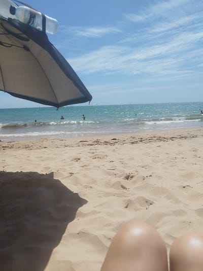 The image captures a serene beach scene in Rota, Spain, on a bright, sunny day. The foreground is dominated by the soft, tan sand of the beach, with gentle undulations and scattered footprints suggesting recent activity. A large beach umbrella, partially obscuring the view, is set up to provide shade. Attached to the umbrella's frame is a clear plastic bottle of water, with a label visible that reads "Morel".

Beyond the sand, the turquoise waters of the sea stretch out to the horizon. Small waves lap gently at the shore, creating a foamy white line where they meet the sand. Several people are visible in the water, either swimming or wading, enjoying the refreshing sea. In the distance, numerous sailboats dot the horizon, adding to the picturesque coastal view.

The sky above is a brilliant blue, punctuated by wispy white clouds that stretch across the expanse, indicative of fair weather. The overall impression is one of a relaxed and pleasant day at the beach, with people unwinding and enjoying the natural beauty of the Spanish coast. The lighting suggests it is likely midday, with the sun high in the sky.