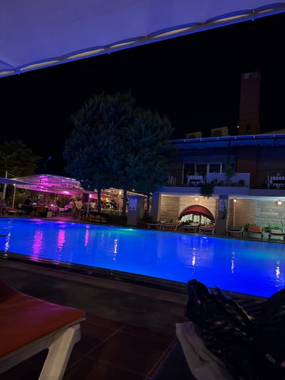 An outdoor swimming pool is illuminated by bright blue lights at night, reflecting the vibrant glow onto the dark surfaces surrounding it. The water has a smooth, glassy appearance, with subtle ripples distorting the reflections of distant lights.

To the left of the pool, an open-air dining area is visible, covered by a white canopy illuminated by pink and purple lights. Several tables are set up, with groups of people seated, engaged in conversation and dining. The atmosphere appears lively and social.

In the background, a multi-story building is visible, constructed with stone and brick. It features balconies with tables and chairs, suggesting it might be a hotel or restaurant. A tall, brick chimney stands prominently on the right side of the building.

The surrounding vegetation consists of dark, leafy trees, providing a contrast to the illuminated pool and dining area. The night sky is dark, with no visible stars or moon.

The foreground of the image shows a portion of a lounge chair, partially covered by what appears to be black plastic bags. The lounge chair has an orange cushion.

The scene is set in Ayvalık, Turkey, likely at a hotel or resort complex.
