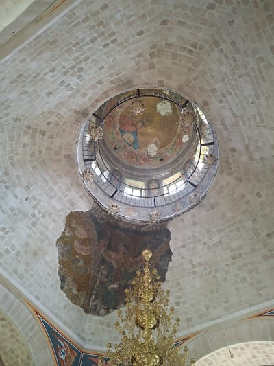 The image shows the interior of a domed structure, likely a church, in Condrița, Moldova. The dome is constructed of light-colored, rectangular stones arranged in a pattern. A circular opening at the apex of the dome is visible, with a metal railing and windows. Below the opening, a circular fresco depicting religious figures in colorful robes is partially visible. Several ornate, gold chandeliers hang from the ceiling. A large section of plaster on the dome has fallen away, revealing a fresco underneath with figures in earthy tones. The frescoed section that remains intact depicts a seated figure in green robes, with a scroll in hand, in front of a decorative border. The overall impression is one of historical architecture with significant artistic and decorative elements, some of which show signs of deterioration.