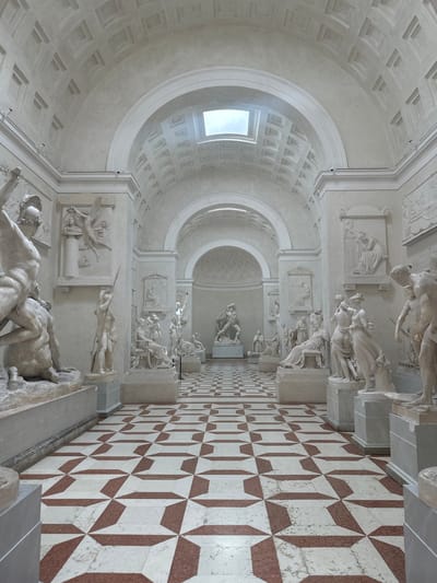 The image depicts a grand, neoclassical-style museum gallery with high, arched ceilings and a skylight. The walls are adorned with bas-relief sculptures and niches containing statues. The floor is paved with a striking geometric pattern of white and reddish-brown marble tiles. Several white marble sculptures of human figures, rendered in classical poses, are displayed throughout the gallery. The overall impression is one of stately elegance and artistic preservation. The location is identified as the Museo Archeologico Nazionale di Cannezza, Cannezza, Italy. There are no people visible in the image.