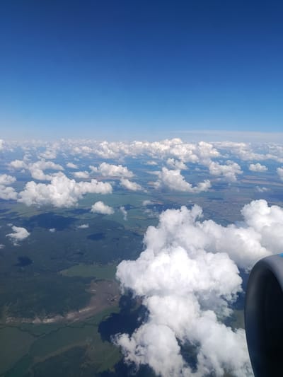 The image is an aerial view of the landscape over Teplovka, Russia, on a clear day with a bright blue sky. Fluffy white clouds are scattered across the sky, casting shadows on the land below. The terrain features a mix of dark green forests and lighter green agricultural fields. In the foreground, a portion of an airplane engine is visible on the right side of the frame. The landscape appears rural and undeveloped, with no signs of urban structures or human activity on the ground. The perspective suggests the photo was taken from an airplane in flight.