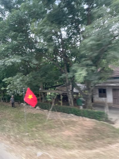 The image, taken at Thôn 18, Vietnam, shows a blurred outdoor scene. In the center is a Vietnamese flag, a red rectangle with a yellow star, attached to a pole. Behind the flag are dense trees with a building partially visible amongst them. A person is standing near the base of the flag. The ground is covered in dirt and grass. The overall image is blurred.