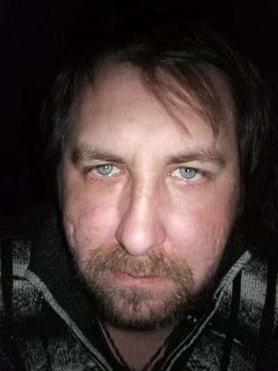 A close-up portrait of a middle-aged man with light blue eyes, light-colored hair, and a beard. He is wearing a dark and light-colored patterned sweater. The background is dark and indistinct. There is no evidence to suggest that this image was taken in Parabel, Russia. No city or location information is possible to determine from this image.
