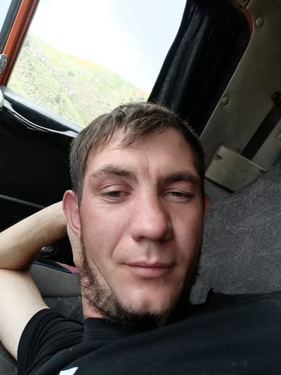 The image is a selfie taken from inside a vehicle. A man with brown hair, a light beard, and a black t-shirt is positioned in the foreground, his head tilted and his left arm bent behind his head. The background shows the interior of the vehicle, including seats and a window through which a green, hilly landscape is visible. The overall lighting suggests it is daytime. The location is identified as Nurekata, Uzbekistan.