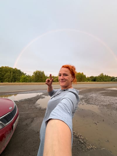 A woman with bright red hair takes a selfie outdoors. She is wearing a light blue long-sleeved shirt and is smiling, holding up two fingers in a peace sign. In the background, a faint rainbow is visible in the sky, suggesting a recent or ongoing rain shower. The ground is wet and muddy, with puddles reflecting the overcast sky. Trees line a road in the middle ground, and part of a red car is visible in the foreground. The scene appears to be in Sovkhoznyi, Russia, possibly during the late afternoon or early evening given the soft light. The overall mood is cheerful and optimistic, despite the damp weather.