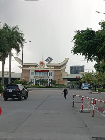 The image shows the Moc Bai border crossing in Ben Cau, Vietnam. A black SUV is parked on the left side of the road, facing away from the camera. In the center of the image, a man in a dark shirt and pants walks towards the border crossing. A white and blue bus is visible in the background, parked on the right side of the road. The border crossing itself is a large, modern building with a distinctive curved roof. The sky is overcast, and the overall lighting suggests it is daytime. Palm trees and other greenery are visible on either side of the road. Red and white striped barriers are positioned along the side of the road.
