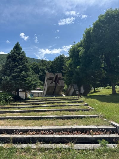 This image captures an outdoor memorial complex or cemetery in Vetren Dol, Bulgaria, under a clear, sunny sky. The scene is static, devoid of people or ongoing activity.
In the midground, a prominent brutalist-style concrete monument stands out, featuring a large, deeply carved five-pointed star on its front face. This central monument is flanked on its right by a series of concrete steps or terraces that extend into the foreground. These steps are interspersed with strips of green grass, some appearing well-maintained, while others contain dried, light brown foliage.
Behind the main star monument, several other rectangular concrete structures are visible, likely grave markers or smaller memorial stones. One of these smaller structures appears to have engraved text, possibly in Cyrillic, with discernible numbers that look like "194...", suggesting a historical date.
The background is dominated by a dense array of green trees, including both broadleaf and coniferous varieties, indicating a forested area. Beyond these trees, a mountain range, also covered in forest, rises under a bright blue sky scattered with fluffy white cumulus clouds. The clear skies and visible shadows suggest it is daytime, likely morning or early afternoon. The overall setting conveys a sense of solemnity and historical remembrance, typical of war memorials or significant commemorative sites.