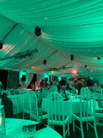 The image captures an indoor evening event within a large tent or marquee, described as being located in Orizari, Bulgaria. The tent's ceiling is draped with white fabric and illuminated predominantly by a vivid green ambient light from multiple spotlights. Numerous strands of warm white fairy lights are strung across the entire draped ceiling. Black spotlights and speakers are mounted to the tent's internal support structures.

The space is furnished with multiple round tables, each covered with a white tablecloth. Each table setting includes several drinking glasses, glass bottles (some containing clear liquid), dark-colored napkins, and floral centerpieces featuring dark flowers and green foliage. White-painted wooden chairs with dark seat cushions are arranged around the tables. In the foreground, a dark suit jacket is draped over one of these chairs.

Approximately 30 to 50 individuals are visible throughout the tent. A significant number of people are congregated in a central area, engaged in dancing and socializing, with some having their arms raised. One person in this central group appears to be holding a microphone or similar device. Other individuals are seated at tables, observing or interacting. The attire of the attendees ranges from semi-formal to formal. Along the tent's sides, white drapes conceal the exterior, and exposed wooden beams wrapped with green vines and leaves are visible, from which a few bare light bulbs hang. Additional stage lighting in magenta and amber hues is present in the central activity area. The floor appears to be a dark, solid surface.