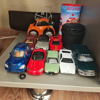 A collection of toy cars is arranged on a gray shelf. The cars are of various colors and models, including a blue PT Cruiser, a red Viper, a green Izh, a white BMW, and a dark green Ram pickup. Behind the cars are an orange toy car with the number 23, a black bag, a red and blue cylindrical container labeled "Let's Play #Kidsattheheart", and a black container with a lid. A small black tire rests near the blue PT Cruiser. The location of the scene is not discernible, nor is there evidence to suggest this is in the city of Pokrovskoye, Russia.
