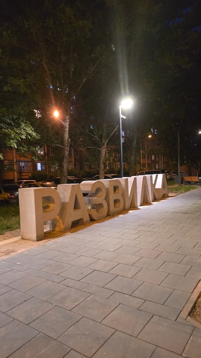 This is a medium shot, nighttime photograph of large, three-dimensional concrete letters spelling "РАЗВИЛКА" (Razvilka) on a paved walkway. The letters are light grey and are illuminated by streetlights. Behind the letters, trees with dark green foliage are visible, as well as apartment buildings with illuminated windows. Several cars are parked along the street in front of the buildings. A lamppost with a bright white light stands to the right of the letters. The walkway is made of grey paving stones laid in a brick pattern. A park bench can be seen in the distance to the right of the letters. The overall scene suggests an outdoor urban environment at night.
