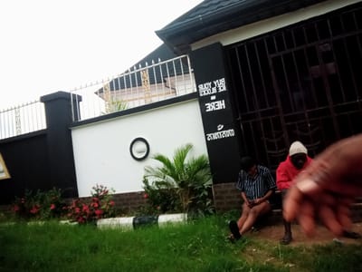 In Nanka, Nigeria, an outdoor scene unfolds with two individuals seated on concrete steps in front of a building. The building features a dark, grated metal gate and a white wall with black accents. Text is visible on the black portion of the wall, appearing to be an advertisement. A young person in a striped shirt and shorts is seated on the left, while another person wearing a red hooded sweatshirt and a white beanie sits to the right. The ground is covered in green grass, and decorative plants with red flowers are visible in front of the building. The sky above is overcast.